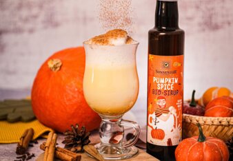 Am Bild ist ein herbstlicher Latte mit Pumpkin Spice Sirup zu sehen, der in ein bauchiges Glas gefüllt wurde und inmitten von Kürbissen steht. Daneben steht eine Flasche Pumpkin Spice Sirup und es rieselt Staubzucker über alles.  | © SONNENTOR