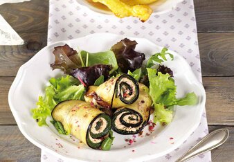 Auf dem Foto sind Melanzani-Röllchen mit Blattsalat auf einem weißen Teller zu sehen. Dahinter steht ein weiterer Teller mit Kartoffelchips. | © SONNENTOR