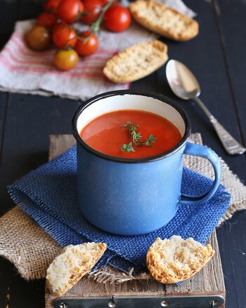 Auf dem Foto sieht man eine Tasse mit Tomatensuppe. Daneben sieht man Baguettes und frische Tomaten. | © SONNENTOR