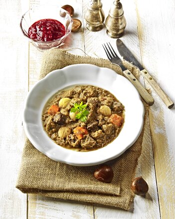 Auf dem Teller sieht man Rehragout mit Maroni. Daneben steht ein Schälchen mit Preiselbeeren. Auch vier ganze Maroni sind neben dem Teller zu sehen. | © SONNENTOR