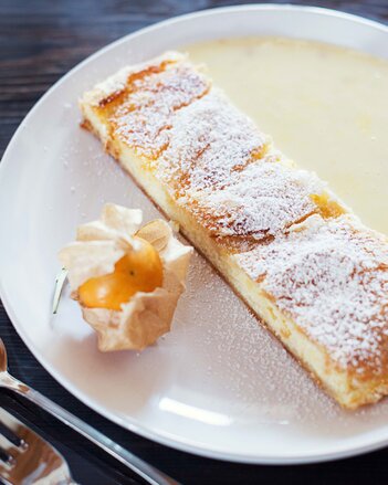 Auf dem Foto ist ein Joghurt-Topfen-Strudel zu sehen. Daneben ist Vanillesauce auf dem Teller zu sehen. Der Strudel ist mit Staubzucker bestäubt. | © SONNENTOR