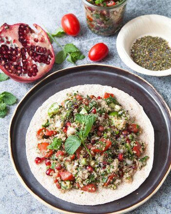 Auf dem Foto sieht man ein Fladenbrot mit Nanas Quinoa Tabouleh darauf. Dahinter sieht man Gewürze, Tomaten und Granatapfel. | © SONNENTOR