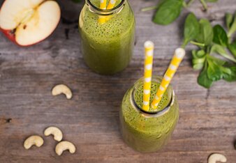 Auf dem Foto sind zwei Flaschen mit Grüner Detox Smoothie zu sehen. Daneben sind Äpfel und Cashewnüsse zu sehen. | © SONNENTOR