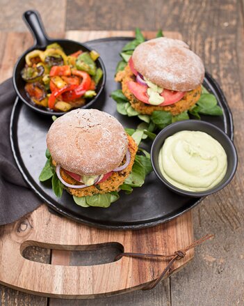 Auf dem Foto ist eine Pfanne mit zwei Holy Veggie Burgern zu sehe. Darauf sieht man auch eine Schale mit Dip und eine Schale mit buntem Gemüse. | © SONNENTOR