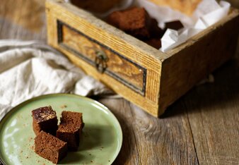 Auf dem Foto sind drei Stück Brownies auf einem grünen Teller zu sehen. Dahinter steht ein Kistchen mit mehreren Brownies darin. | © SONNENTOR