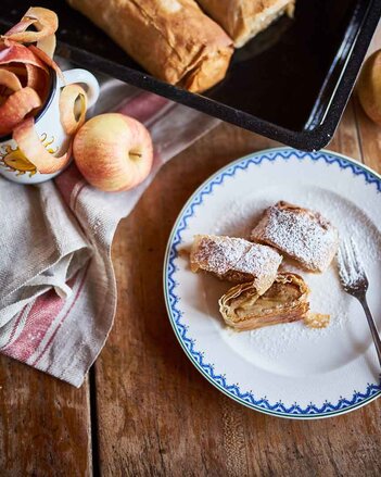 Auf dem Foto ist ein Teller mit Apfelstrudel zu sehen. Er ist mit Staubzucker bestäubt. Dahinter steht ein schwarzes Bleich mit zwei Apfelstrudel.  | © SONNENTOR