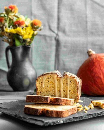 Auf dem Foto ist ein aufgeschnittenes Kürbisbrot mit Karamellglasur zu sehen. Im Hintergrund steht eine Blumenvase und ein Kürbis. | © SONNENTOR