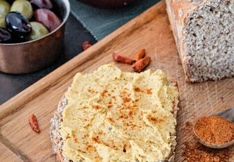 Ein Sonnenblumenbrot mit Harissa Aufstrich liegt auf einem Brett neben Chilischoten und dem Harissa Gewürz, das in einem Löffel ist. In einer Schale daneben befinden sich schwarze und grüne Oliven. | © SONNENTOR