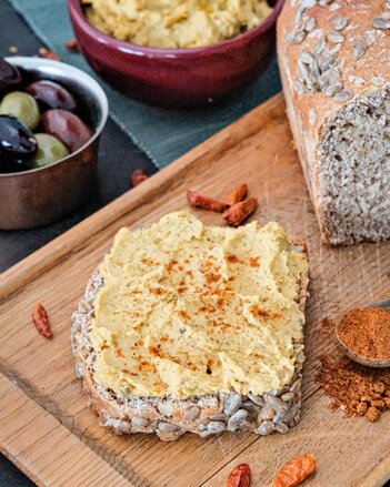 Ein Sonnenblumenbrot mit Harissa Aufstrich liegt auf einem Brett neben Chilischoten und dem Harissa Gewürz, das in einem Löffel ist. In einer Schale daneben befinden sich schwarze und grüne Oliven. | © SONNENTOR