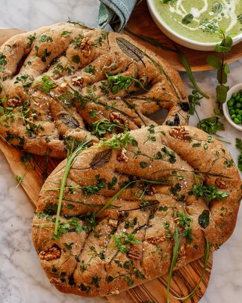 Zwei Stück Brot-Gebäck mit regelmäßigen Einschnitten, belegt mit Walnüssen und Wildkräutern, die fast aussehen, wie ein Zopf, liegen auf einem Holzbrett. | © SONNENTOR