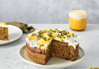 Die Pumpkin Spice Torte mit einer weißen Creme mit Kürbiskernen und Pumpkin Spice Sirup darauf ist bereits angeschnitten und steht auf einer Tortenplatte. Im Hintergrund steht ein Glas mit Pumpkin Spice Latte.  | © SONNENTOR