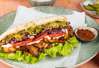 Der Tofu Kebab ist von oben geschichtet gefüllt mit grünem Paprika, roten Zwiebeln, Tomatenscheiben, Sauce, Tofu und einem Salatblatt und liegt auf einem Teller neben einem Schüsselchen Harissa-Gewürz. Im Hintergrund steht eine weiße Dip-Sauce in einem Schälchen und man sieht 2 Paradeiser.  | © SONNENTOR