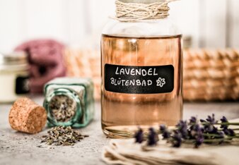 Auf dem Foto ist ein Fläschchen mit Lavendelblütenbad zu sehen. Davor sieht man den violetten Lavendel und Kräuter. | © SONNENTOR