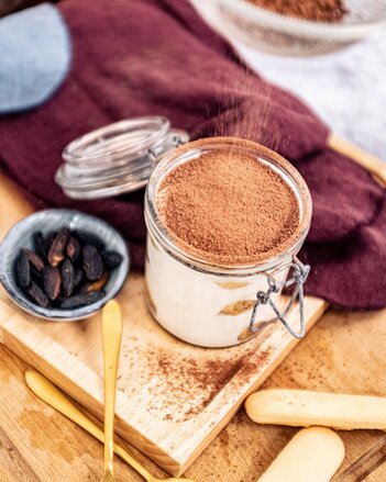 Auf dem Foto sieht man ein Glas mit Tiramisu. Auf das Tiramisu wird Zimt gestreut. Daneben sieht man eine Schüssel mit Tonkabohnen. | © SONNENTOR