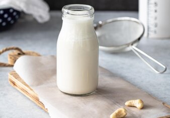 Glasflasche mit pflanzlicher Sahne steht auf einem Holzbrett. Darum herum ein paar Cashewnüsse. | © SONNENTOR