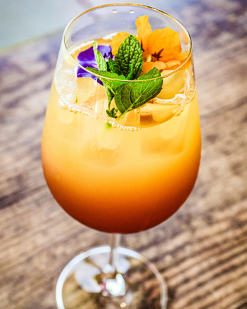 Der orangefarbene Cocktail ist in ein schönes Weinglas gefüllt, unten ist er dunkler als oben. Es befinden sich Eiswürfel mit im Glas und alles ist mit essbaren Blüten und Minzblättern dekoriert. | © SONNENTOR