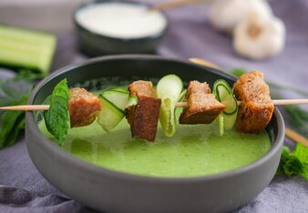 The cucumber soup is served in a black soup bowl, decorated with a skewer with rolled cucumber strips, bread croutons and mint leaves. | © SONNENTOR