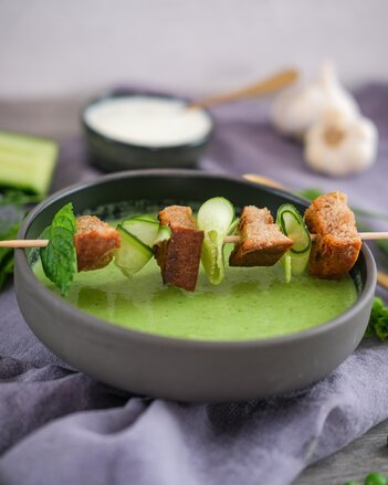 The cucumber soup is served in a black soup bowl, decorated with a skewer with rolled cucumber strips, bread croutons and mint leaves. | © SONNENTOR