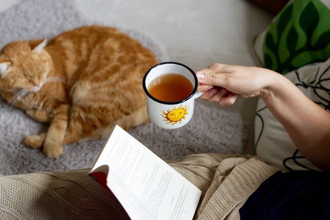 Auf dem Foto trinkt eine Frau auf dem Sofa eine Tasse Tee und liest ein Buch. Daneben sieht man eine Katze liegen. | © SONNENTOR